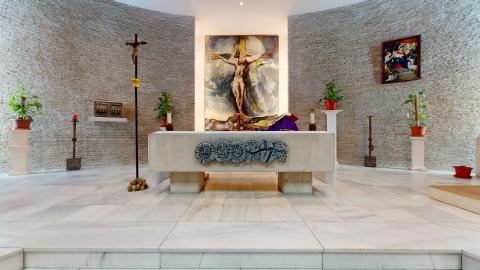 Capilla. Espacio sagrado de ambiente sereno y luminoso con paredes curvas revestidas de piedra gris texturizada y suelo de mármol blanco. En el centro destaca un altar de piedra clara con un relieve tallado de peces. Al fondo, preside la sala un gran cuadro luminoso de la crucifixión de Jesús. La estancia está decorada con varias plantas verdes sobre pedestales blancos.