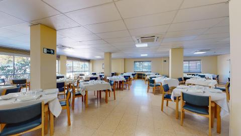 Comedor. Amplio comedor distribuido en una sala diáfana con columnas cuadradas de color crema y suelo de terrazo claro. Cuenta con múltiples mesas cuadradas vestidas con manteles blancos y preparadas con vajilla. Las sillas son de madera con tapizado azul oscuro. Al fondo y a los lados hay amplios ventanales con persianas de lamas que aportan luz natural.