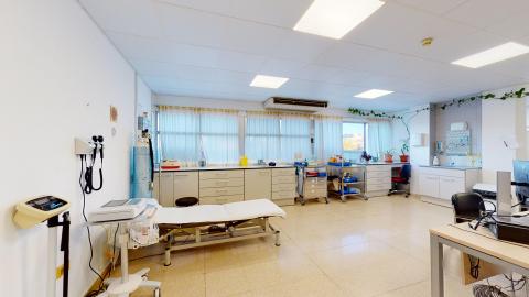 Consulta de enfermería. Consulta médica espaciosa y luminosa con paredes blancas y suelo de terrazo claro. A lo largo del fondo hay un gran ventanal cubierto con cortinas finas y un mueble blanco con cajoneras. En primer plano destaca una camilla de exploración blanca y, a la izquierda, una báscula médica y un tensiómetro de pared. Una enredadera decora la parte superior de las ventanas.