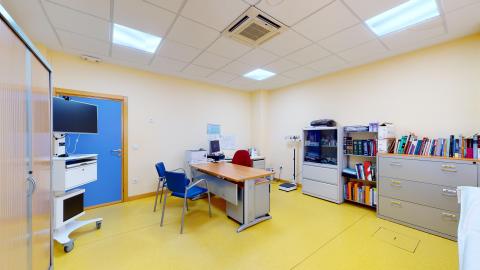 Consulta médica. Despacho médico de tonos cálidos con paredes de color amarillo claro y suelo de vinilo amarillo intenso. En el centro hay una mesa de escritorio de madera con una silla de oficina roja y dos sillas azules para confidentes. A la derecha destaca una estantería metálica repleta de libros y un archivador gris. Una puerta azul resalta a la izquierda.