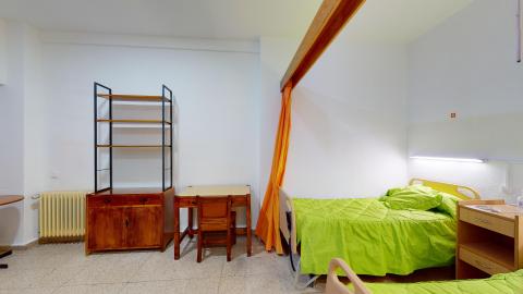 Habitación doble. Habitación doble de paredes blancas y suelo de terrazo. Cuenta con dos camas articuladas vestidas con una colcha de color verde lima llamativo. Una cortina de color naranja cuelga de una viga de madera en el techo para separar los ambientes. El mobiliario incluye una mesilla de noche, un escritorio con silla y una estantería alta, todos de madera oscura.