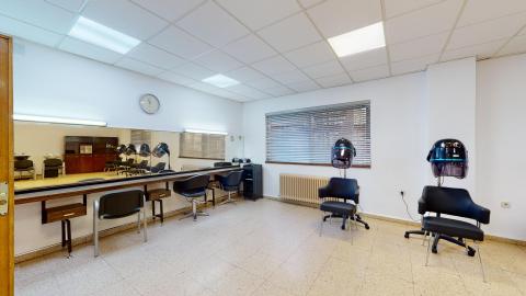 Peluquería. Sala de peluquería amplia y muy luminosa con paredes blancas y suelo de terrazo claro. Destaca un gran espejo frontal iluminado con tres sillones negros de estética frente a él. A la derecha, junto a un amplio ventanal con persianas, hay otros dos sillones equipados con secadores de casco negros independientes.