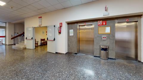 Ascensores. istribuidor interior con suelo de terrazo oscuro y brillante. Destacan dos ascensores contiguos con puertas de acero inoxidable encastrados en una pared de cemento gris. A la izquierda hay un extintor de incendios colgado en la pared blanca y unas puertas dobles abiertas que muestran una zona de espera con sillas de madera.