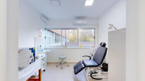 Consulta de podología. Consulta médica de podología pequeña, ordenada y con abundante luz natural gracias a un amplio ventanal al fondo que da al exterior. La sala tiene paredes y mobiliario blancos. Destaca en el centro un sillón clínico articulado de color negro, acompañado de un taburete con ruedas y diverso equipamiento médico a los lados.