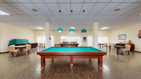 Sala de actividades. Sala de ocio amplia con paredes de tono amarillo claro y suelo de terrazo oscuro. En el centro destacan dos mesas de billar de madera y tapete verde, iluminadas por tres lámparas de techo con pantallas de color verde intenso. Alrededor hay distribuidas varias mesas cuadradadas con sillas de madera clara para juegos.