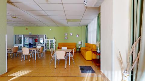 Sala de estar. Sala de descanso muy luminosa con grandes ventanales cubiertos por cortinas verdes y suelo de baldosas amarillas. Cuenta con una pared de acento pintada en verde pistacho frente a la cual destacan dos llamativos sofás de color amarillo mostaza. En el centro hay varias mesas de madera clara con sillas a juego.