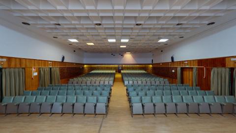 Salón de actos. Gran salón de actos de ambiente sobrio con un techo técnico cuadriculado de color blanco y paredes revestidas de paneles de madera oscura. Destacan numerosas filas de butacas tapizadas en color verde oliva, dispuestas de forma simétrica a ambos lados de un pasillo central de madera clara que conduce al escenario.