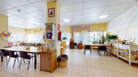 Terapia ocupacional. Taller de manualidades muy amplio y luminoso, rodeado de grandes ventanales con cortinas blancas. Las paredes combinan el blanco y el amarillo, decoradas con murales coloridos y un gran árbol dibujado. La sala está equipada con mesas grandes de trabajo, sillas negras y estanterías llenas de materiales de artesanía y plantas.