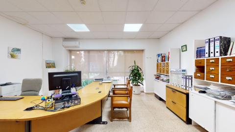 Trabajo social. Oficina de trabajo social de tonos neutros, con paredes blancas y suelo de terrazo claro. Dispone de un gran ventanal al fondo con estores beige que iluminan la estancia. Destaca una gran mesa de madera curvada con un ordenador en primer plano, flanqueada por sillas de cuero marrón y varias estanterías blancas repletas de archivadores de oficina.