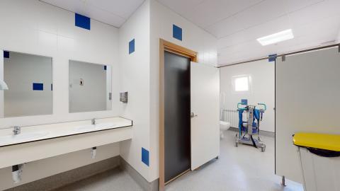 Baño. Un baño geriátrico amplio, con paredes de azulejos blancos y detalles azules. Tiene dos lavabos integrados en una encimera blanca, dos espejos encima, y un WC con barras de apoyo. La sala está bien iluminada por la luz que entra por una ventana y luces en el techo. En el centro, destaca una grúa móvil de color azul para el traslado de personas.
