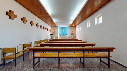 Capilla. Una capilla con paredes blancas y bancos de madera. El techo tiene detalles de madera y luces integradas, además de luz natural que entra por varias ventanas pequeñas en el lateral derecho. Al fondo, un altar con un mantel blanco y, detrás, una pared turquesa con un gran crucifijo de madera. En las paredes laterales, hay catorce cruces de madera del Vía Crucis.