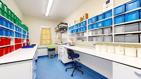 Depósito de medicamentos. Un depósito de medicamentos con estanterías blancas y suelo azul. Paredes blancas y mucha iluminación por fluorescentes en el techo. El espacio está repleto de contenedores de plástico de colores (rojo, azul y verde) para organizar los fármacos sobre mesas blancas. Hay un escritorio con una silla azul y, al fondo, una ventana con cortinas amarillas.