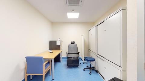 Consulta de podología. Una consulta de podología con paredes blancas y suelo azul. En el centro, destaca un sillón de podología negro con reposacabezas y reposabrazos, flanqueado por un taburete giratorio azul. A la izquierda, una mesa de oficina con una silla azul. A la derecha, armarios blancos grandes con puertas correderas. La iluminación es intensa y proviene del techo.