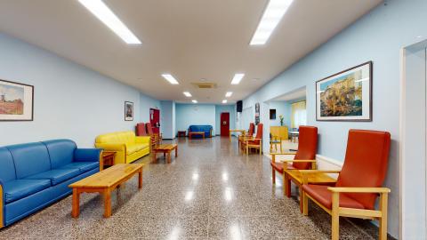Sala de estar. Vista amplia de una sala de estar muy bien iluminada con luz artificial en el techo y paredes de tono azul claro. El suelo es de terrazo pulido con motas. A ambos lados del pasillo central se disponen sillones tapizados en colores azul, amarillo y rojo, junto con mesas bajas de madera de pino. Al fondo se vislumbran puertas y cuadros en las paredes.