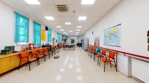 Terapia ocupacional. Una gran sala rectangular con abundante luz artificial blanca. Paredes blancas decoradas con un gran mapamundi y estanterías de madera con diversos materiales de terapia. En el centro, una larga hilera de sillas naranjas y turquesas con estructura negra se alinea a lo largo de un pasillo central. Al fondo hay mesas cuadradas y armarios de almacenamiento. El suelo es de baldosas claras.