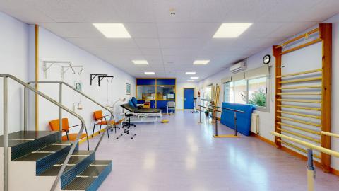 Fisioterapia. Un gimnasio amplio y luminoso, con luz natural que entra por una gran ventana lateral. El suelo es de linóleo lila y las paredes de un tono suave a juego. Incluye equipos de fisioterapia como espalderas de madera, una escalera con barandilla en primer plano, colchonetas azules enrolladas, y una camilla de tratamiento al fondo. El espacio se siente limpio y diáfano.