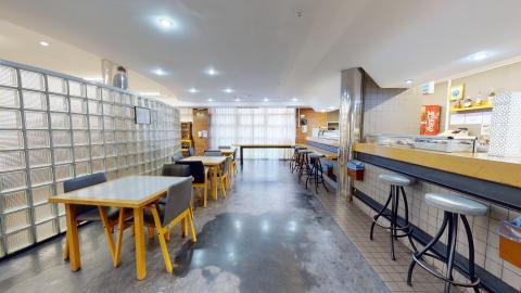 Cafetería. Cafetería iluminada con luz blanca artificial y natural al fondo a través de unas cortinas claras. Destaca un muro curvo de bloques de vidrio traslúcido a la izquierda que separa unas mesas de madera con sillas oscuras. A la derecha hay una barra de bar larga con taburetes altos de metal y asientos grises sobre un suelo oscuro brillante.
