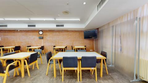 Cafetería. Sala con paredes de ladrillo visto de tono arcilloso y gran luminosidad proporcionada por tubos fluorescentes en el techo. Dispone de varias mesas de madera con sillas tapizadas en negro. Destaca a la derecha un panel divisor de plástico traslúcido junto a un gran ventanal cubierto por una cortina de color beige.