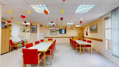 Centro de día. Sala de estar amplia y muy luminosa con paredes de color crema y suelo de baldosas grises. Al fondo a la derecha se observa un gran ventanal con cortinas azuladas. Destaca la decoración festiva de otoño con hojas de papel de colores colgadas del techo y dos grandes mesas con llamativas sillas de color rojo intenso.