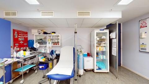 Consulta de enfermería. Consulta médica de paredes blancas y suelo gris con una iluminación artificial muy clara. En el centro destaca una camilla de exploración con base azul y tapicería blanca. Alrededor se distribuyen un armario acristalado con medicamentos, un carro de curas metálico y un refrigerador blanco para vacunas.