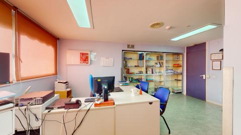 Consulta médica. Despacho médico con paredes de color violeta claro y un ventanal a la izquierda cubierto por un estor naranja que tamiza la luz natural. Destaca una mesa de escritorio beige en ángulo con un ordenador, rodeada de sillas de oficina de color azul vivo. Al fondo hay una gran estantería acristalada llena de libros.