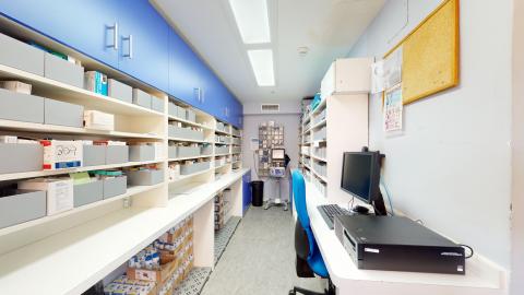 Depósito de medicamentos. Habitación estrecha y alargada destinada a farmacia, con paredes blancas y excelente iluminación cenital blanca. A la izquierda destaca una larga bancada blanca bajo una fila de armarios superiores azules, con múltiples cajetines organizadores de medicamentos grises. A la derecha hay una mesa con un ordenador y silla azul.