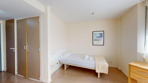 Habitación. Dormitorio acogedor con paredes blancas y suelo de tono tostado, iluminado por una ventana lateral fuera de cuadro. Destaca una cama individual con ropa de cama blanca y una manta beige. Sobre la cama cuelga un cuadro. Se ven a la izquierda un armario empotrado y la puerta del baño y del acceso.