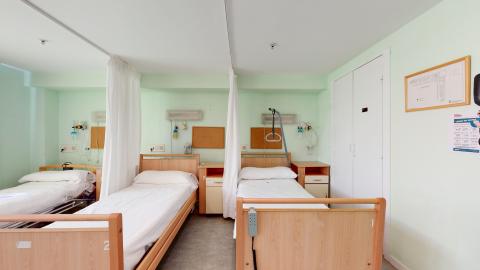 Habitación de enfermería. Habitación de enfermería con tres camas articuladas separadas por cortinas. Cuenta con equipamiento médico en la pared de la cabecera, mesillas de noche y una ventana con vistas al exterior. Las paredes son de color claro.