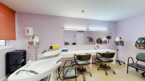 Peluquería. Sala de peluquería con paredes de color lila suave, un gran espejo, lavabos con sillas negras y secadores de casco. Cuenta con una ventana que aporta luz natural.