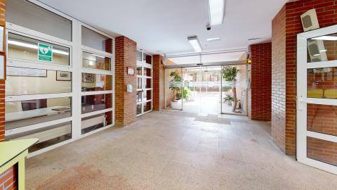 Vestíbulo. Entrada a una sala con grandes ventanales que dan al patio exterior. Al fondo se ve una zona común con más ventanas y vegetación. Las paredes son de ladrillo visto y blanco.