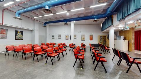 Salón de actos. Salón de actos espacioso con suelo de cemento pulido gris y paredes blancas. El techo alto tiene conductos de aire azules expuestos y luces fluorescentes. Varias filas de sillas tapizadas en rojo brillante están organizadas mirando hacia un escenario al fondo, delimitado por cortinas rojas. Destaca la presencia de varios pósters artísticos enmarcados colgando de las paredes.