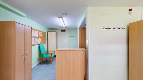 Enfermería. Zona de control de enfermería con iluminación artificial de tubos fluorescentes. Paredes verde menta claro y suelo azul. Un mostrador de madera clara domina el primer plano, con el letrero "CONTROL ENFERMERIA" visible en la pared. Al fondo, se ven armarios de madera y un sillón reclinable turquesa. Pasillo con puertas y un extintor rojo en la pared.