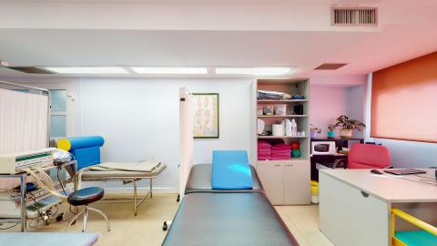 Fisioterapia. Sala de fisioterapia dividida por cortinas blancas en cubículos. Bien iluminada por luces de techo y una ventana lateral con estor naranja. Paredes azul lavanda y suelo gris. Cuenta con varias camillas de tratamiento (una negra con cojín azul), un escritorio blanco con silla rosa, estanterías con suministros y equipos médicos variados sobre carritos.