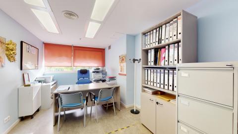 Trabajo social. espacho luminoso con paredes azul cielo y suelo gris. Cuenta con una gran ventana con estor naranja que aporta luz natural. En el centro, un escritorio con dos sillas de oficina azules para visitantes. Una estantería alta llena de archivadores blancos y un archivador de metal beige ocupan la pared derecha. Un perchero blanco está en el rincón.