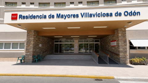 Acceso. Entrada principal exterior del edificio bajo una luz diurna intensa y despejada. Destaca una gran marquesina blanca con letras azules donde se lee "Residencia de Mayores Villaviciosa de Odón", flanqueada por columnas revestidas de piedra marrón. En la zona de acceso, de tonos neutros y grisáceos, se aprecian puertas acristaladas, una rampa de accesibilidad con barandilla metálica, ventanas laterales y varias sillas de plástico verde.