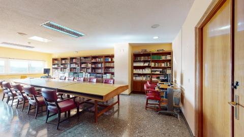 Vista general de una biblioteca espaciosa y bien iluminada por luz natural que entra por un gran ventanal al fondo. Predominan las estanterías de madera repletas de libros, una larga mesa central con sillas de color granate y, a la derecha, puestos individuales con ordenadores. Las paredes son de tono beige claro y el suelo es de terrazo oscuro.