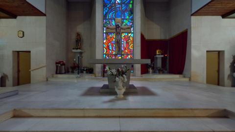 Capilla. Imagen interior del altar de una capilla moderna y luminosa. El elemento más destacado es una gran vidriera central de colores intensos con un crucifijo en el medio. Delante, el altar de piedra sobre un podio elevado. El espacio está revestido con grandes losas de piedra blanca y paneles de madera en el techo. A la izquierda hay una figura religiosa y a la derecha unas cortinas rojas.