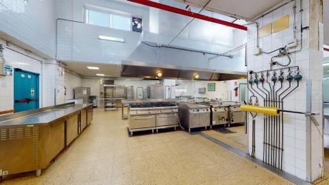 Cocina. Cocina industrial amplia, limpia y bien iluminada. Destaca el equipamiento profesional de acero inoxidable, incluyendo grandes fogones, hornos y encimeras. En primer plano, las instalaciones de tuberías y grifería de gas están visibles en una pared de azulejos blancos. El suelo es de baldosas de color beige. Se perciben varias ventanas altas que aportan luz natural.