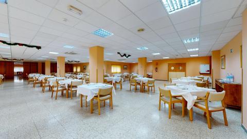 Comedor. Vista general de un amplio y luminoso comedor preparado con numerosas mesas redondas vestidas con manteles blancos y sillas de madera clara. Las paredes son de un tono melocotón cálido y el suelo es de terrazo claro. Varias columnas estructurales cuadradas están pintadas del mismo color que las paredes. El techo es de placas con iluminación fluorescente integrada. Se aprecian algunas ventanas al fondo.