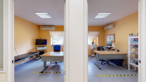 Consultas médicas. Fotografía de dos despachos médicos contiguos e idénticos, separados por un panel central blanco, vistos desde la entrada. Ambos tienen paredes de color melocotón, suelos de baldosas grises y ventanas con cortinas blancas al fondo. Cada espacio cuenta con un escritorio de madera, una silla de oficina azul y equipos médicos. En el despacho de la izquierda hay una camilla y un televisor; en el de la derecha, un monitor y un armario vitrina. Marcas amarillas y negras delimitando espacios en el suelo.