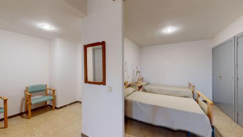 Habitación doble. Habitación doble limpia y sencilla, dividida parcialmente por un tabique central que alberga un espejo. Las paredes son blancas y el suelo es de terrazo claro. A la derecha, se ven dos camas individuales paralelas vestidas con colchas beige. A la izquierda, un par de sillas de madera con tapicería azul claro están dispuestas para visitas. Dos luces de techo cuadradas iluminan la estancia. Al fondo, unas puertas grises.