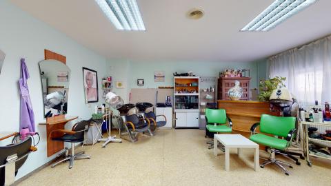 Peluquería. Sala de peluquería luminosa con paredes de color verde agua muy claro y suelo de terrazo amarillento. Cuenta con varios puestos de trabajo provistos de espejos, sillones negros y secadores de casco profesionales. En la zona de espera destacan dos llamativos sillones de cuero verde vivo junto a una pequeña mesa baja blanca, iluminados por paneles fluorescentes en el techo y una ventana al fondo con cortinas blancas.