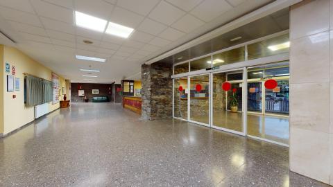 Recepción. Amplio pasillo y zona de entrada visto desde el interior, con un suelo gris moteado muy brillante que refleja la luz de los paneles del techo. A la derecha destaca el gran frente acristalado de la puerta de salida, señalizada con un cartel de «SALIDA» y círculos rojos de seguridad en los cristales. A la izquierda se aprecian paredes de color amarillo claro y un mostrador de recepción al fondo.