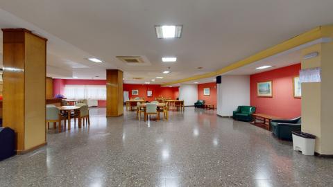Sala de actividades. Gran sala multiusos con suelo de terrazo gris y paredes que combinan el color blanco con un llamativo tono rojo arcilla al fondo. El espacio está iluminado por plafones rectangulares en el techo y cuenta con varias mesas de comedor de madera rodeadas de sillas. Destaca a la derecha una zona de descanso con confortables sillones de cuero verde oscuro alineados junto a mesas auxiliares.