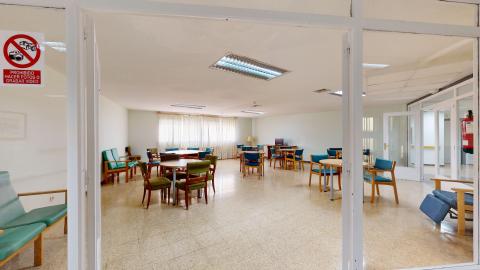Sala de estar. Sala de estar amplia y muy luminosa fotografiada desde el marco de una puerta blanca. La estancia dispone de suelo claro y paredes blancas, con luz natural que proviene de un gran ventanal al fondo cubierto por cortinas finas. El espacio está distribuido con múltiples mesas redondas y cuadradas acompañadas de sillas con tapicería azul y verde, además de varios sillones de descanso orejeros.