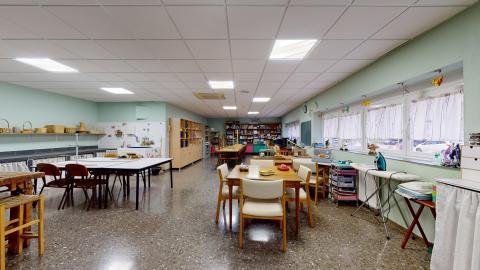 Terapia ocupacional. Extensa sala de terapia ocupacional con paredes pintadas en verde menta claro y suelo de terrazo oscuro. Dispone de una iluminación excelente gracias a los paneles del techo y a una hilera de ventanas con cortinas cortas a la derecha. Destacan múltiples mesas de trabajo de madera, estanterías repletas de materiales, máquinas de coser y un área con tablas de planchar y cestas de mimbre.