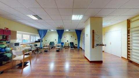 Fisioterapia. Amplia y luminosa sala de fisioterapia con suelo de parqué marrón y paredes de color amarillo claro. A la izquierda, destaca un gran ventanal que aporta luz natural, junto a estanterías con material deportivo como balones medicinales. Al fondo, se disponen varias camillas de tratamiento separadas por cortinas de color azul intenso. En primer plano a la derecha, destaca una gran columna beige con un aparato de ejercicios y una espaldera de madera en la pared lateral.