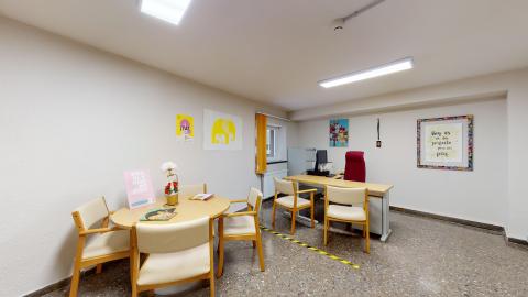 Consulta de psicología. Despacho luminoso con paredes blancas y suelo de terrazo gris. La sala combina dos zonas: en primer plano a la izquierda, una mesa redonda de madera clara con cuatro sillas a juego; al fondo, un escritorio de trabajo con ordenador y una silla de oficina con un llamativo respaldo alto de color granate. El espacio cuenta con una pequeña ventana con cortina amarilla que aporta luz natural y está decorado con dibujos y un cuadro colorido con un mensaje positivo en la pared derecha.