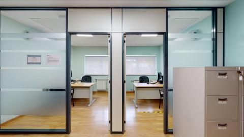 Trabajo social. Vista frontal de dos despachos idénticos y contiguos, separados por mamparas de cristal translúcido con perfiles gris oscuro. Los despachos tienen suelo de madera clara, paredes de un suave tono verde agua y cuentan con una ventana al fondo que ilumina el espacio. Cada habitáculo dispone de una mesa de oficina de madera clara y una silla de ruedas negra. En el extremo inferior derecho, en primer plano fuera de los despachos, destaca un archivador metálico gris de cajones.