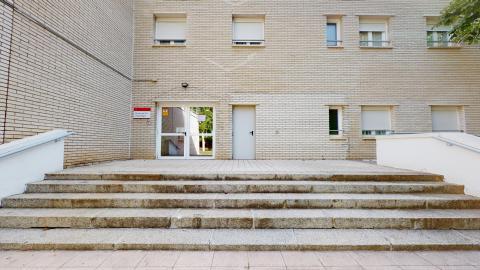 Acceso centro de día. Entrada exterior a nivel de calle con una escalinata de piedra de cinco peldaños que sube hacia una terraza pavimentada. La fachada es de ladrillo visto de tono claro y cuenta con varias ventanas con persianas bajadas. Destaca una puerta doble acristalada con marco blanco bajo un cartel que indica "Centro de Día Vista Alegre", situada junto a otra puerta metálica gris opaca. La iluminación es de luz natural diurna de intensidad media.